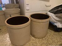 Pair of beige stoneware pickling crocks with dark brown glazed interiors placed side by side on a patterned floor with other crocks visible in the background.