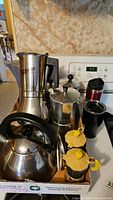 Photo showing a variety of stove top espresso makers, a steel kettle, two yellow plastic containers, and two metallic drinking bottles, one new