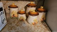 Five vintage mushroom ceramic canisters in varying sizes displayed on countertop. Each has a mushroom cap lid with small mushroom knob and mushroom decorations on body.