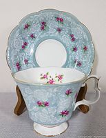 Teacup front with saucer displayed on a wooden stand, showing light blue background, white floral embossing, pink rosebuds, and gold trim.