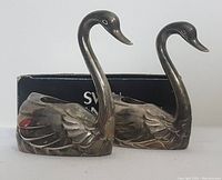 Two silver plated swan-shaped candle holders shown side by side with detailed views of their shape and design, positioned in front of a black box.