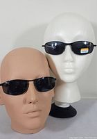 Two pairs of sunglasses displayed on mannequin heads, showing the front view of both pairs with black frames and dark lenses.