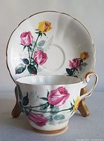 Close-up photo of white bone china teacup and matching saucer with pink and yellow rose floral pattern and gold trim. Teacup positioned in front of saucer.