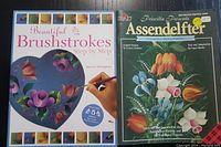 Photo showing two softcover books side by side on a dark surface. Left book titled 'Beautiful Brushstrokes Step by Step' with a heart-shaped palette featuring colorful floral brushstrokes. Right book titled 'Priscilla Presents Essendelefter' featuring traditional Dutch floral painting on cover.