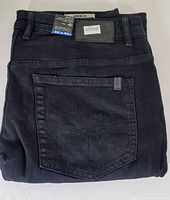 Folded black men's jeans showing back pocket and leather-look patch on waistband with brand tags attached.
