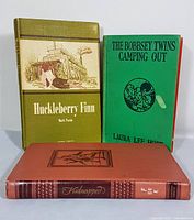 Three vintage hardcover books standing and lying: The Bobbsey Twins Camping Out, Kidnapped, Huckleberry Finn showing covers and spine