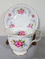 Bone china teacup and saucer with pink rose and grey scroll pattern, gold trim, both displayed against white background with wooden stand for saucer.