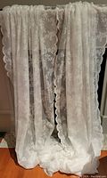 Photo of two white lace window panels hanging, showing their length and floral scalloped edges.