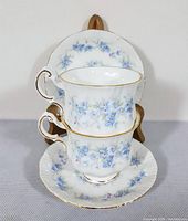 Two bone china cups stacked in front of a matching saucer displaying blue floral pattern and gold trim.