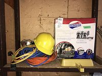 Photo of a shelf with coiled orange and blue extension cords, yellow hard hat, and American Tourister car organizer box marked new.