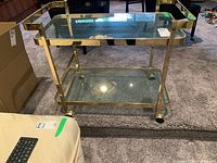 Front view of metal and glass rolling cart with two glass shelves and brass-colored frame on caster wheels.