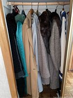 Seven women's coats hanging on a wooden rod including various colors and materials such as denim blue, camel tan, grey, and tweed with fur collars.