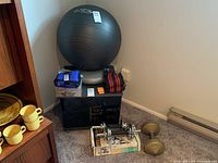 Wide shot including black resistance ball, ankle weights in box, waist weights on top, two sets of dumbbells, balance cushion, all arranged on top and around a small black cabinet.