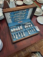 Wooden case containing 1847 Rogers Bros silverplate flatware including forks, spoons, and knives displayed on a table with surrounding china plates.