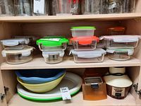 Shelf with various glass food storage containers with plastic lids in different shapes and colors, serving bowls and plates below, plus a popcorn maker and a small kitchen tool.