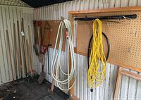 Overview of garden tools including hoes, shovels on the left side, two garden hoses hanging on pegs on the right.