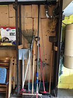 Full view of garden tools propped against a wooden wall, including shovels, brooms, and a rake