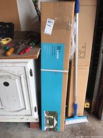 Tall rectangular box with a For Living branded label showing an image of the gazebo setup outdoors beside a broom and some tools.