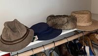 Four men's hats hanging on a clothing rod: brown leather hat, blue wide-brim hat, faux fur winter hat, and straw sun hat.