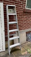 Side view of the six foot aluminum step ladder leaning against a brick wall; the ladder has rubber feet and a built-in paint tray at the top with multiple holes, showing some paint splatters.