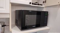 Black Panasonic NN-SG626B microwave oven on a shelf in a kitchen, showing front view with the digital control panel illuminated.
