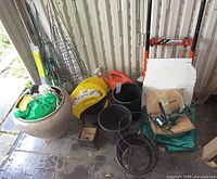 Overview of lot showing ceramic plant pot, various black plastic pots, tomato cages, orange watering cans, leaf scoops, hand truck, and bags of gardening material.