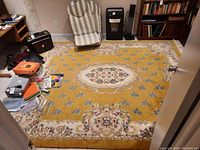 Yellow wool area rug with cream floral medallion and scattered flowers, fringed edges visible, room setting with chairs and assorted items on the rug.