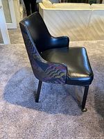 Side view of a chair showing black faux leather seat, armrest, and multi-colored patterned fabric on the side and back, two front legs on casters, two stationary rear legs