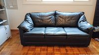 Front view of black leather sofa bed with three seat cushions in a room with parquet floor.