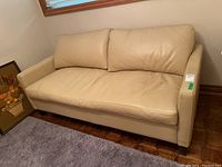 Side angled view of beige leather sofa showing left arm, cushions, and parquet flooring beneath. A framed art piece is partly visible on the left against the wall.
