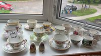 Photo showing various teacups and saucers arranged on windowsill including floral and scenic patterns, also salt and pepper shakers in front.
