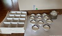 Set of 12 Aynsley teacups and saucers arranged beside a white storage box with 12 compartments.