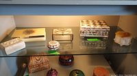 Full view of all eight assorted trinket boxes on glass shelf showing varying sizes, colors, and materials including mother of pearl inlaid boxes, composite flower box, small cream box, cloisonne style pill box, and various rectangular boxes.