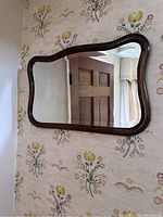 Full view of antique mirror mounted on wall showing curved dark brown wooden frame with floral wallpaper background.