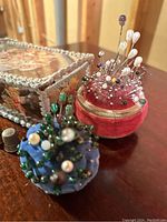 Close up of two velvet pincushions filled with assorted hat pins and sewing pins, next to a floral crocheted clear plastic box and a metal thimble