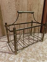 Front and side angled view of brass magazine rack with bamboo-style frame and handle on tiled floor in front of wood panel wall.