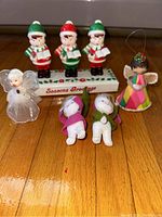 Overall lot showing lighted choir boys display standing on base with Seasons Greetings text, surrounded by small angel and elf ornaments on wood floor.
