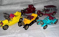 Five vintage die cast car models arranged showing side and front profile, featuring yellow, maroon, gray, and turquoise colors with red and brass details.