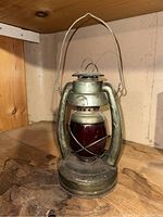 Full frontal photo of metal railroad lantern with red glass globe, standing on wooden surface. Metal handle and red globe wires are clearly visible. Shows wear and patina consistent with age.