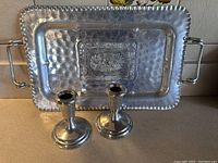 Full view of hammered aluminum tray with embossed center and two metal handles behind pair of pewter candlesticks on a flat surface