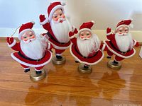 Front view of four Santa Claus figurines with plastic faces, white beards, and felt suits standing on gold bases.