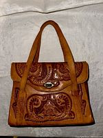 Front view of a hand tooled brown leather Flores handbag, showing floral and swirl tooling, flat leather shoulder straps, and metal twist-lock clasp.