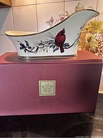 Side angle of Lenox porcelain sleigh-shaped wine caddy on original box, showing cardinal and holly design with gold rims.