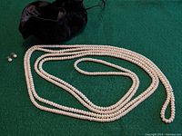 Photo showing long faux pearl necklace arranged on green surface alongside pair of faux pearl stud earrings and small black drawstring pouch.