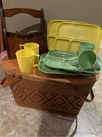 Vintage woven picnic basket with leather handles and diamond pattern, topped with plastic divided trays and mugs in yellow and green, wooden flatware caddy in background.
