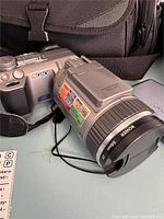 Close front angle view of the Sony DSC-F707 digital camera showing lens with stickers and lens cap, camera body detail, strap.