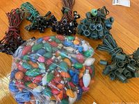Five bundles of vintage light strands with attached plugs and empty bulb sockets, arranged around a plastic bag containing loose large colored bulbs.