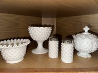 Five white hobnail milk glass pieces displayed on shelf, showing ruffled edge bowl, pedestal compote, salt and pepper shakers with metal tops, and covered sugar dish.