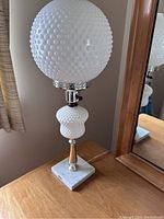 White hobnail glass lamp with round hobnail globe shade on a silver metal stem and square white marble base, shown on wooden dresser beside mirror.