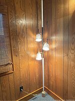 Full view of the white pole lamp standing against wood-paneled wall, showing three lampshades lit and the wooden extension block at the base.